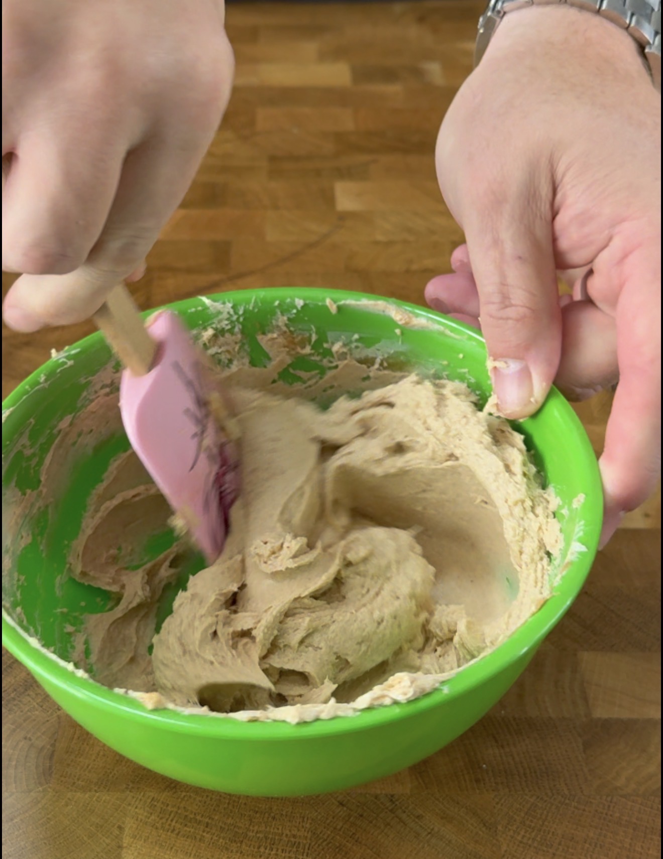 A person mixes cookie dough with a pink spatula in a green bowl on a wooden countertop.