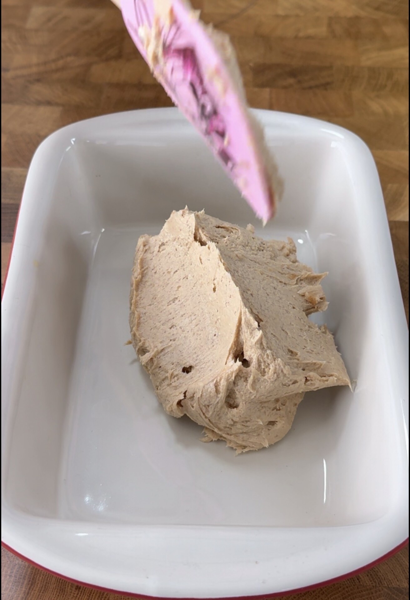 A pink spatula with tan batter over a white rectangular baking dish with a dollop of the same batter inside, sitting on a wooden countertop.