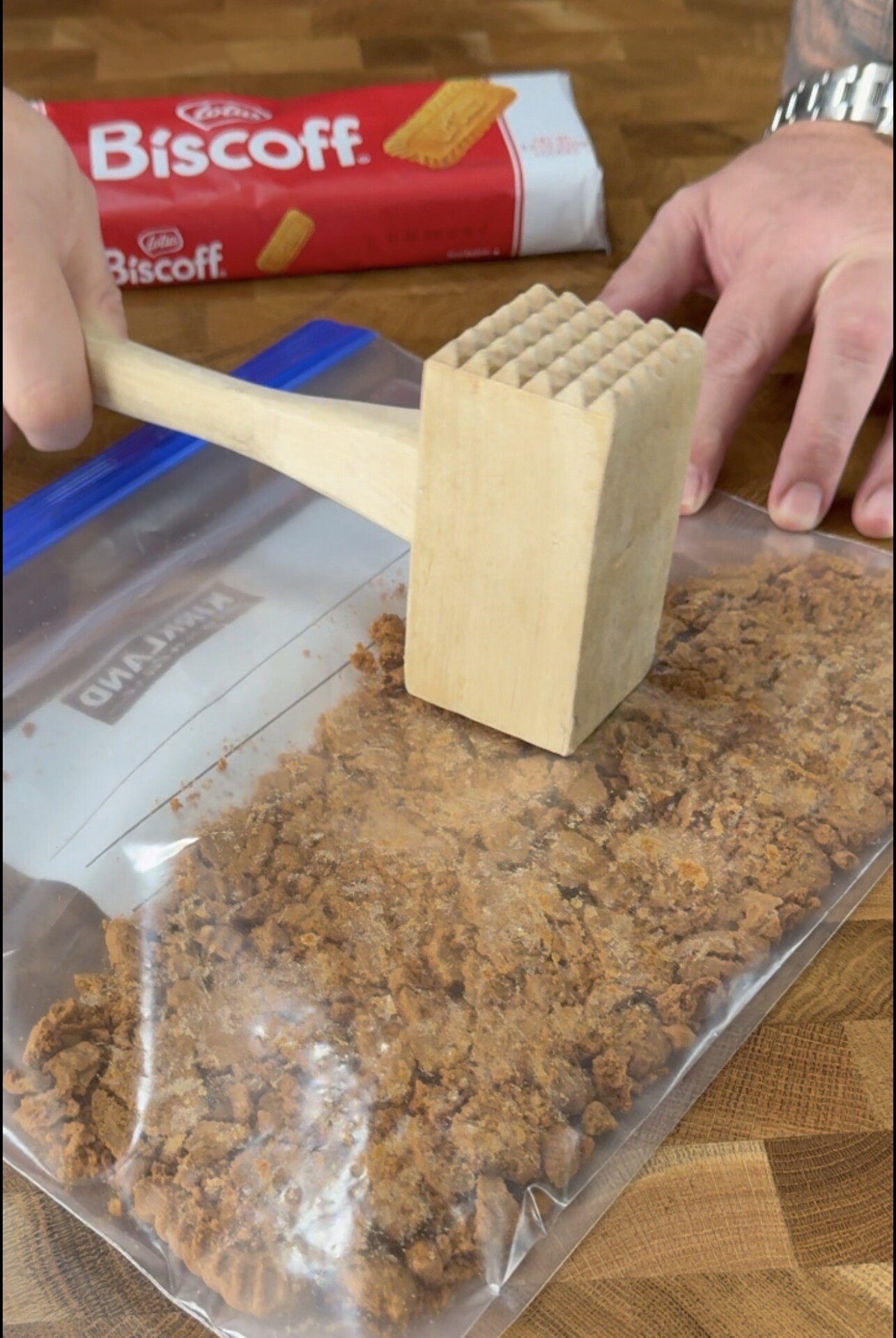 A person crushes Biscoff cookies in a ziplock bag with a wooden mallet on a wooden countertop.