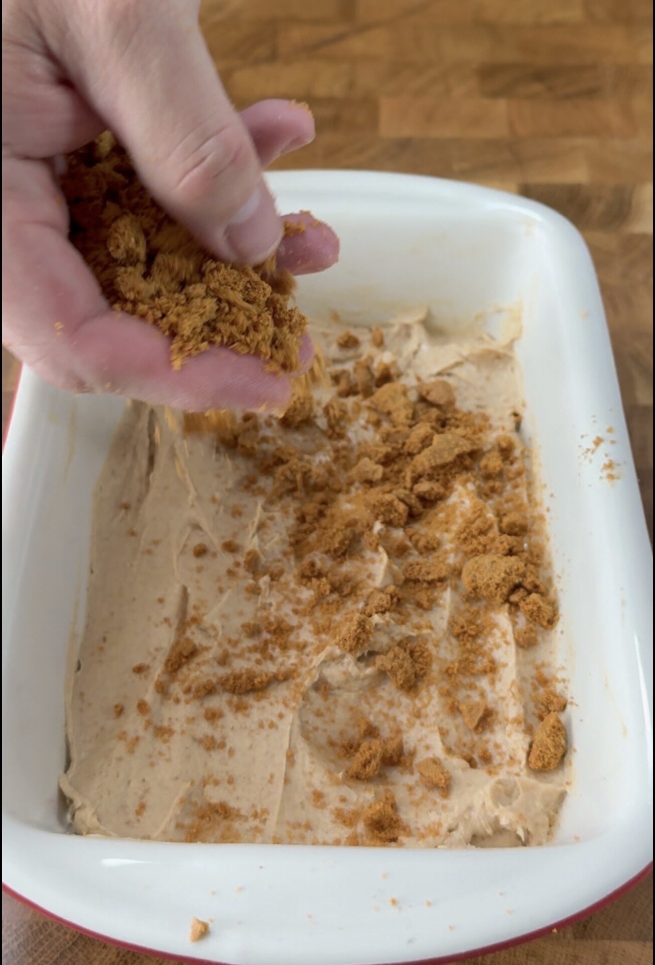 A hand sprinkles crumbled cookies over a creamy layer in a white rectangular dish, set on a wooden surface.