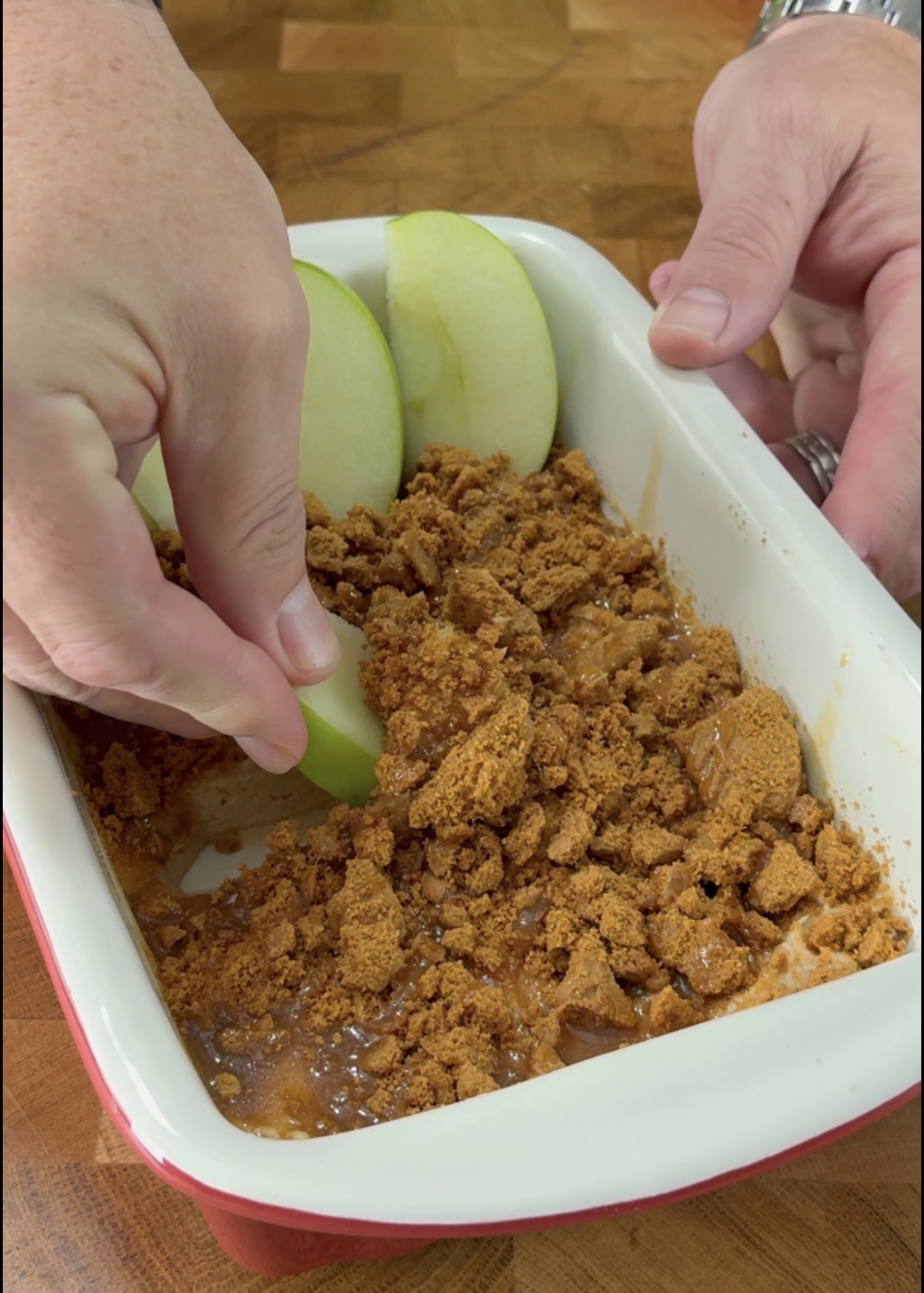 Hands dipping a green apple slice into a rectangular dish filled with crumbled cookies and caramel sauce, on a wooden surface.