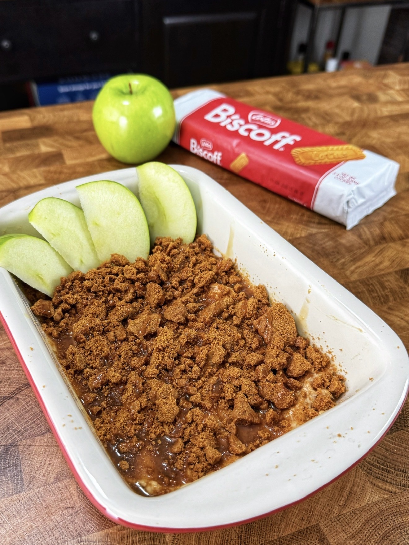 A dish with sliced green apple and crumbled Biscoff cookies on top, a whole green apple beside it, and a Biscoff biscuit package in the background, all on a wooden surface.