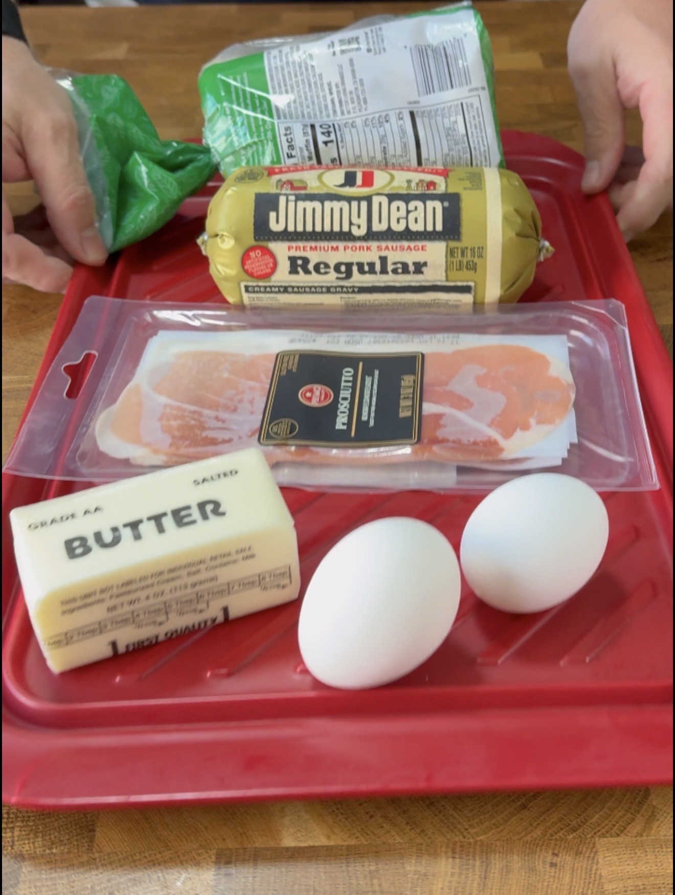 Hands holding a green bag near a red tray with pork sausage, prosciutto, a stick of salted butter, and two white eggs on a wooden surface.