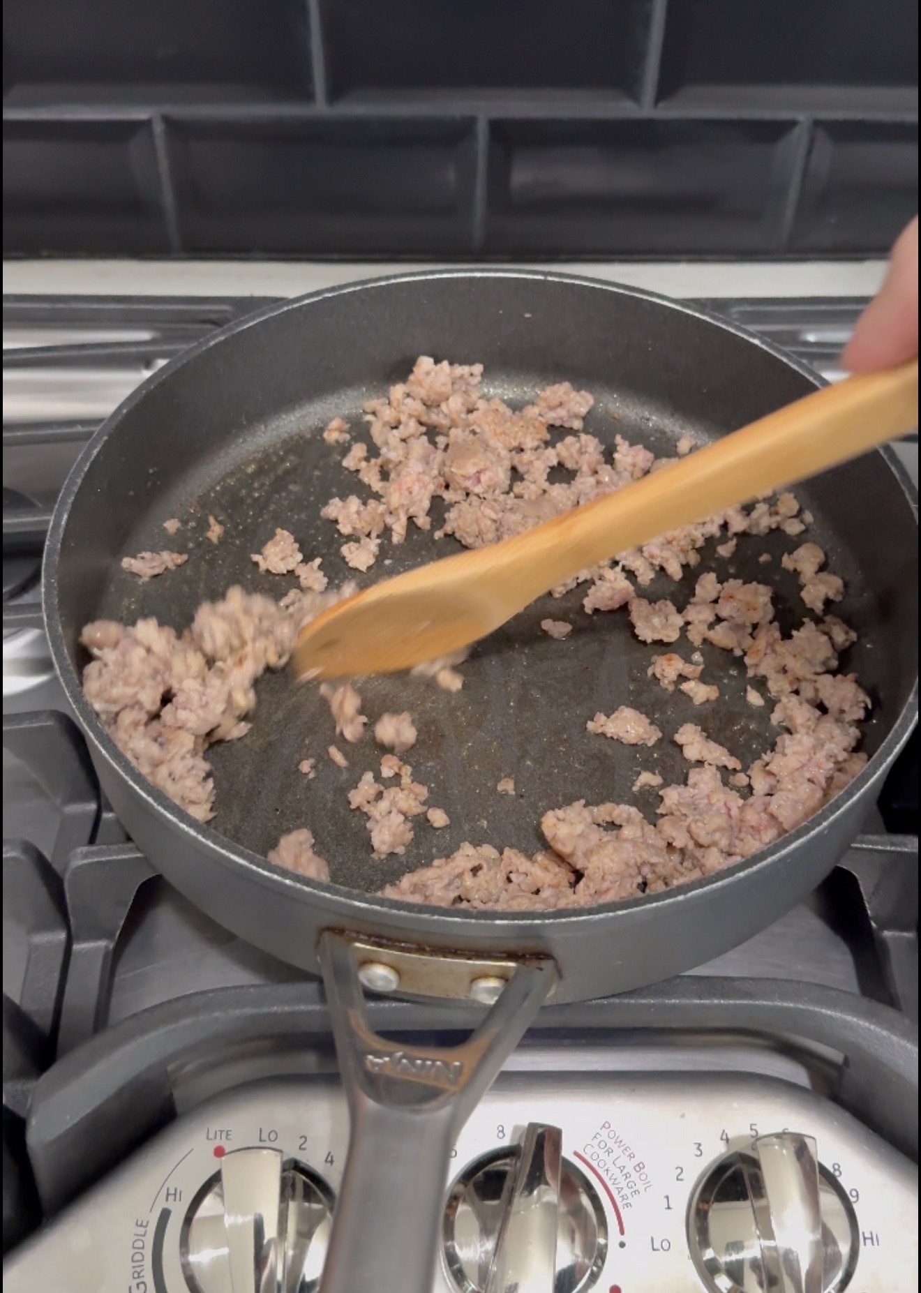 Ground meat is being stirred in a black frying pan on a stove using a wooden spatula. The stove dials are visible at the bottom of the image.