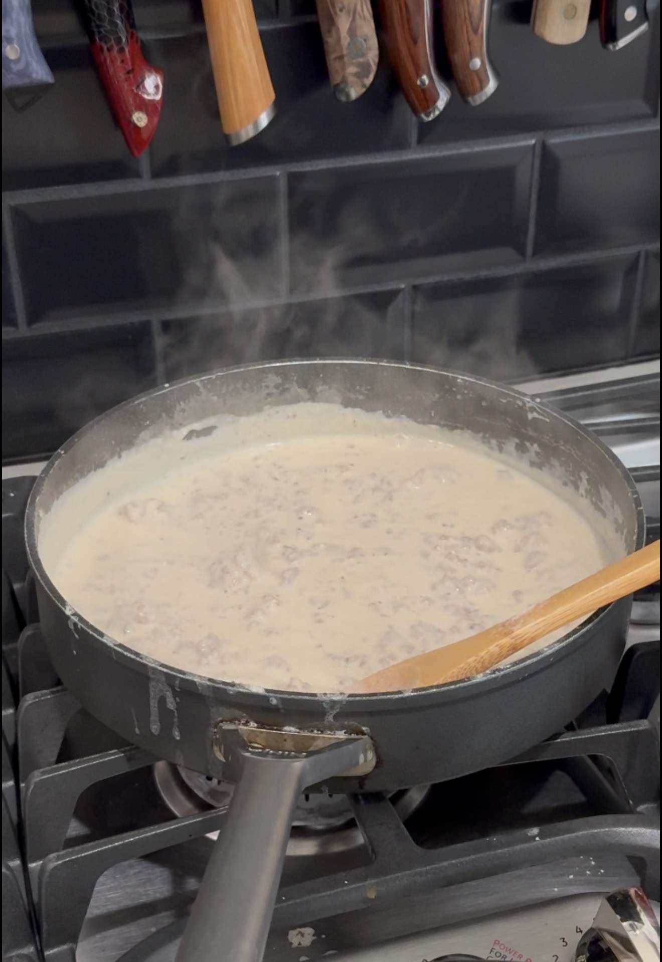 A pan of creamy sausage gravy simmers on a gas stove, steaming, with a wooden spoon resting on the edge. Several kitchen knives hang on a magnetic strip in the background.