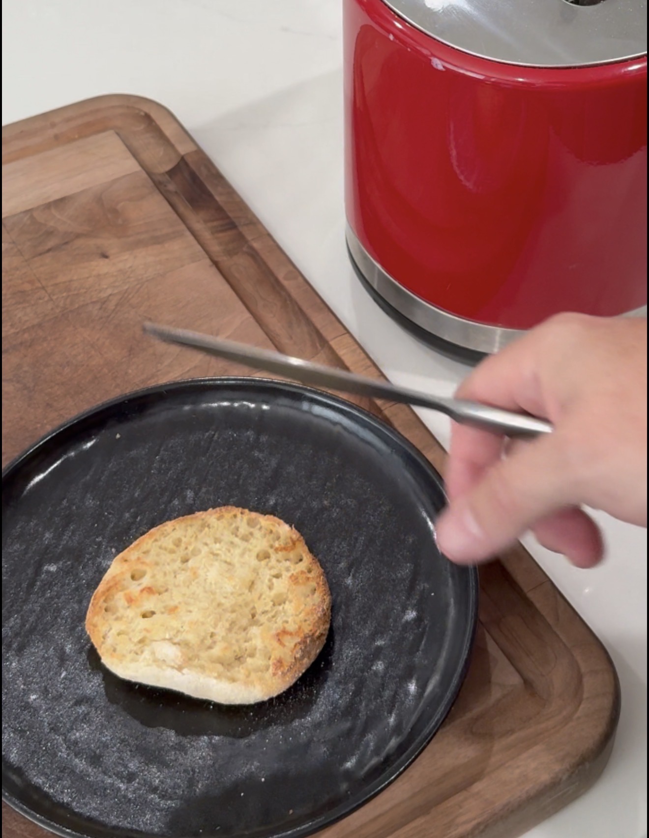 A hand holds a knife above a toasted English muffin half on a black plate, with a wooden cutting board and a red appliance nearby.