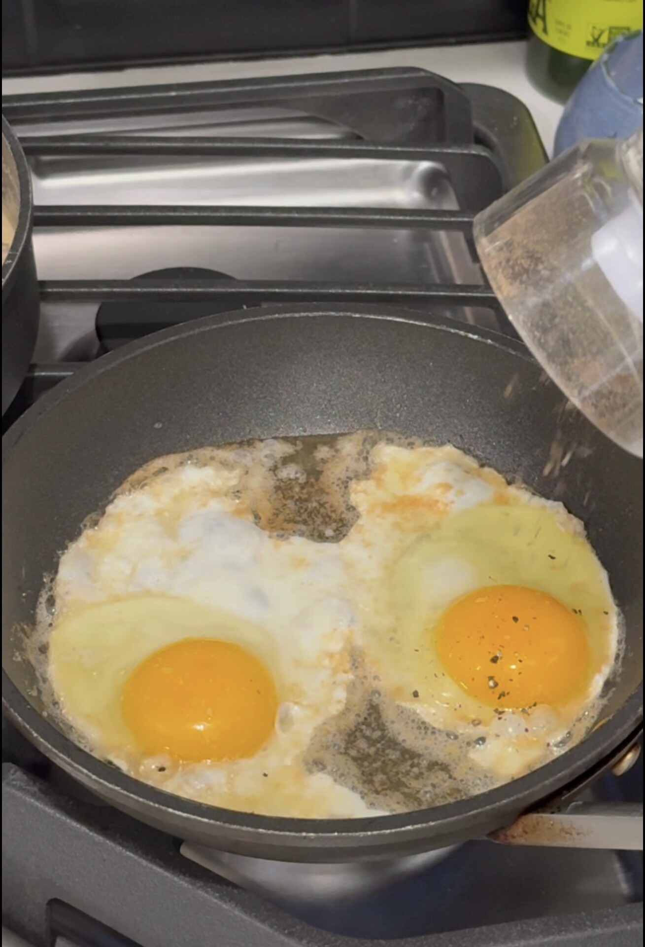 Two sunny-side-up eggs cooking in a nonstick frying pan on a gas stove, with cracked black pepper sprinkled on the eggs. A hand is holding a glass bowl above the pan.