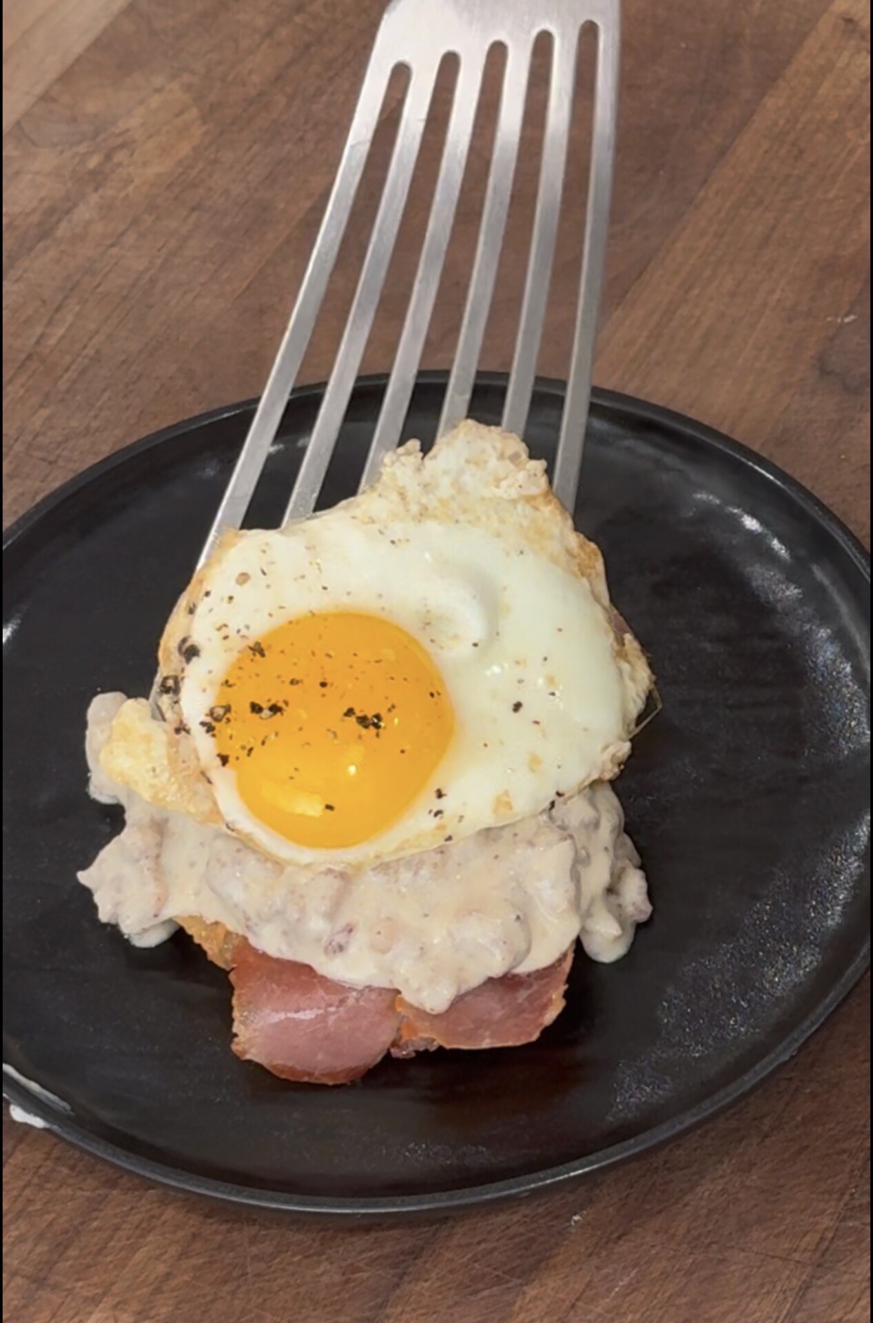 A spatula lifts a stack of toast topped with ham, creamy gravy, and a sunny-side-up egg, all served on a black plate over a wooden table.