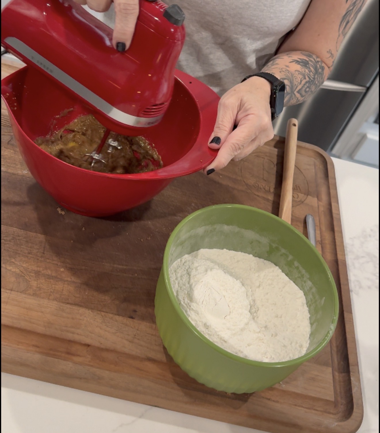 A person uses a red hand mixer to blend ingredients in a red bowl. Next to it is a green bowl filled with flour on a wooden cutting board. The person has tattoos and is wearing a smartwatch.