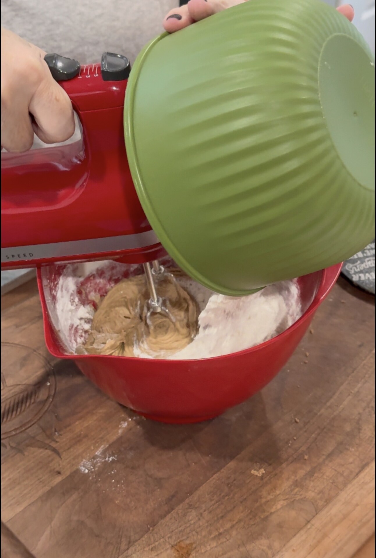 A person pours dough from a green bowl into a red bowl containing flour, using a red hand mixer to combine the ingredients on a wooden surface.