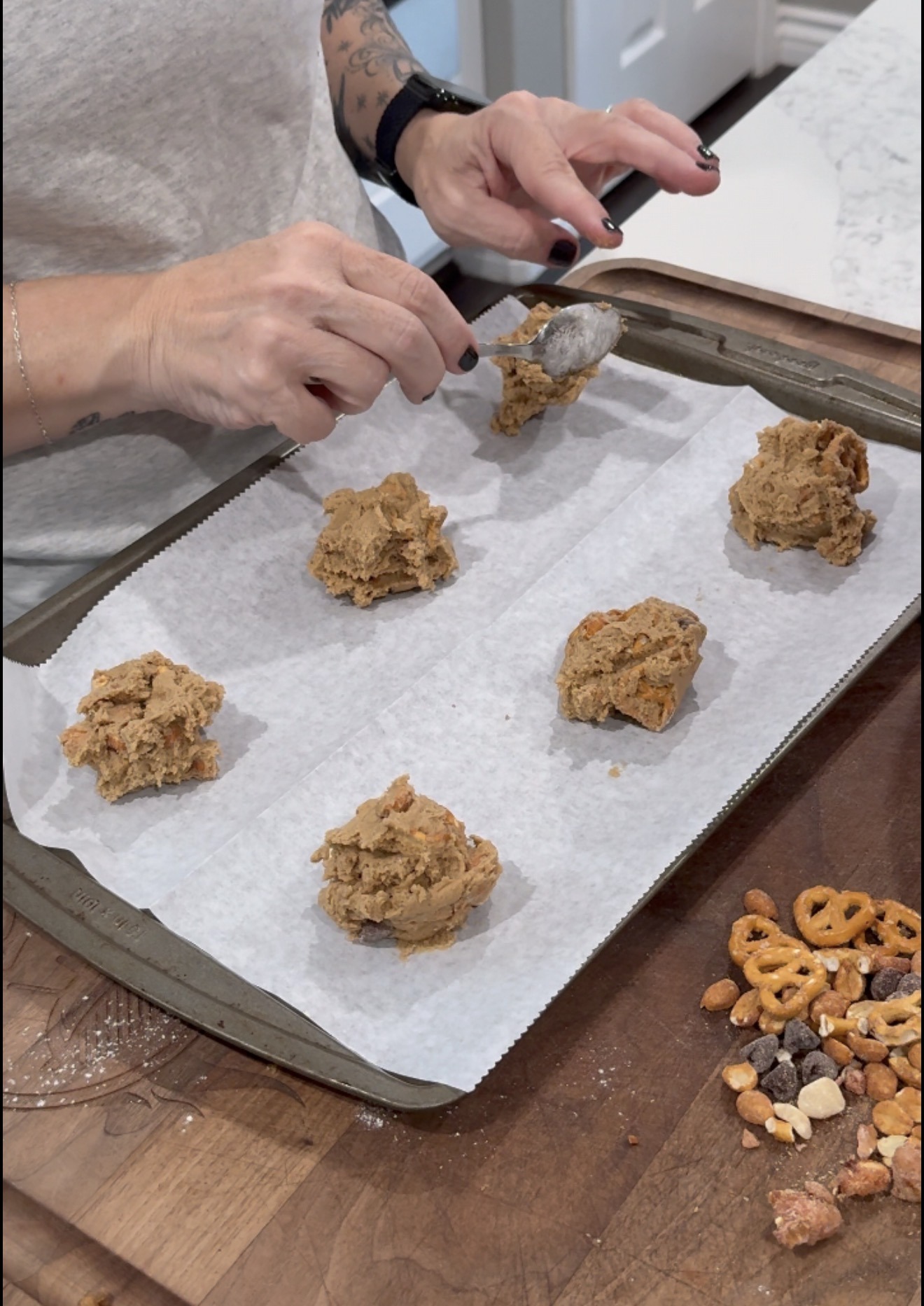 Person scooping cookie dough onto a parchment-lined baking sheet; pieces of pretzels and nuts are visible on the wooden counter nearby.