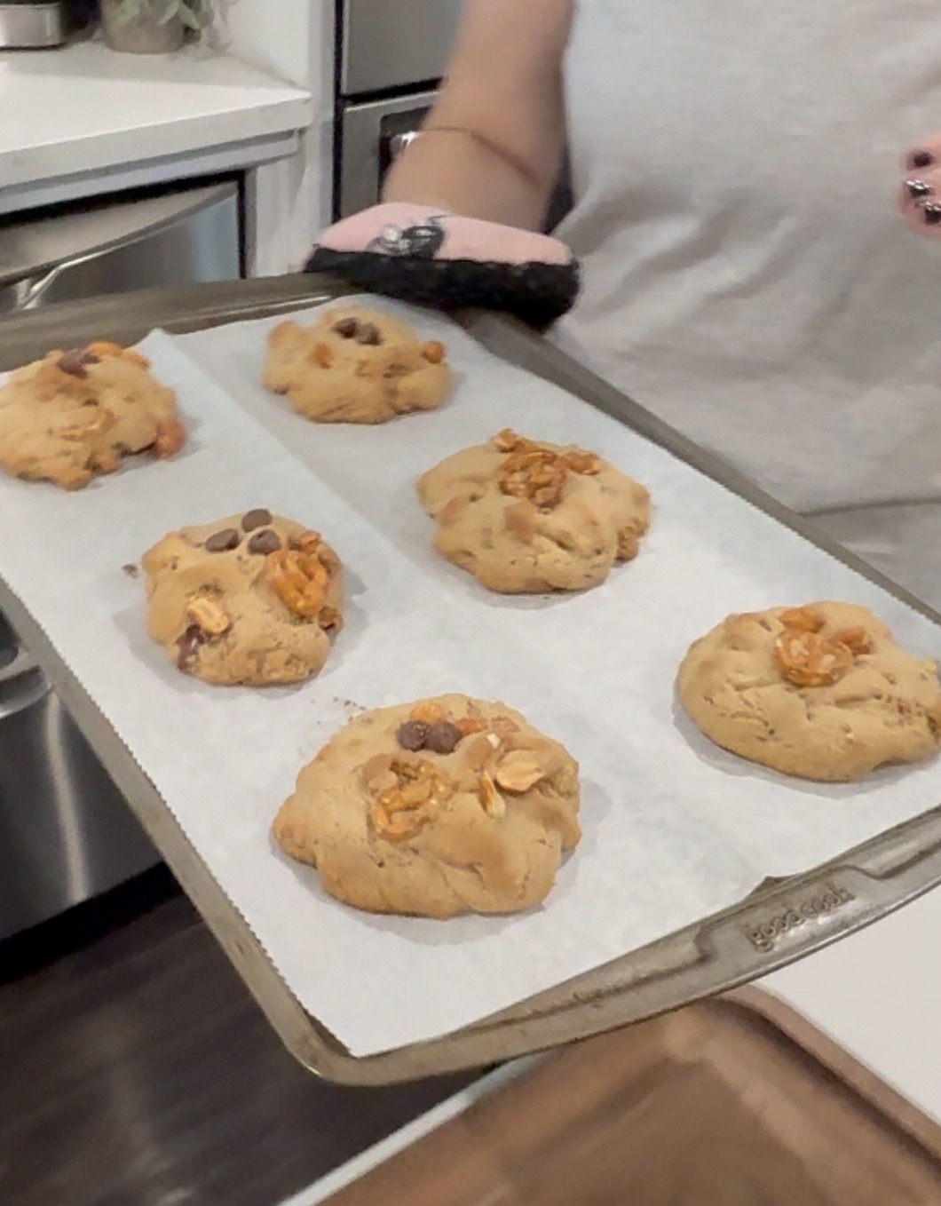 A person holds a baking tray lined with parchment paper, displaying six freshly baked cookies with chocolate chips and nuts in a kitchen.