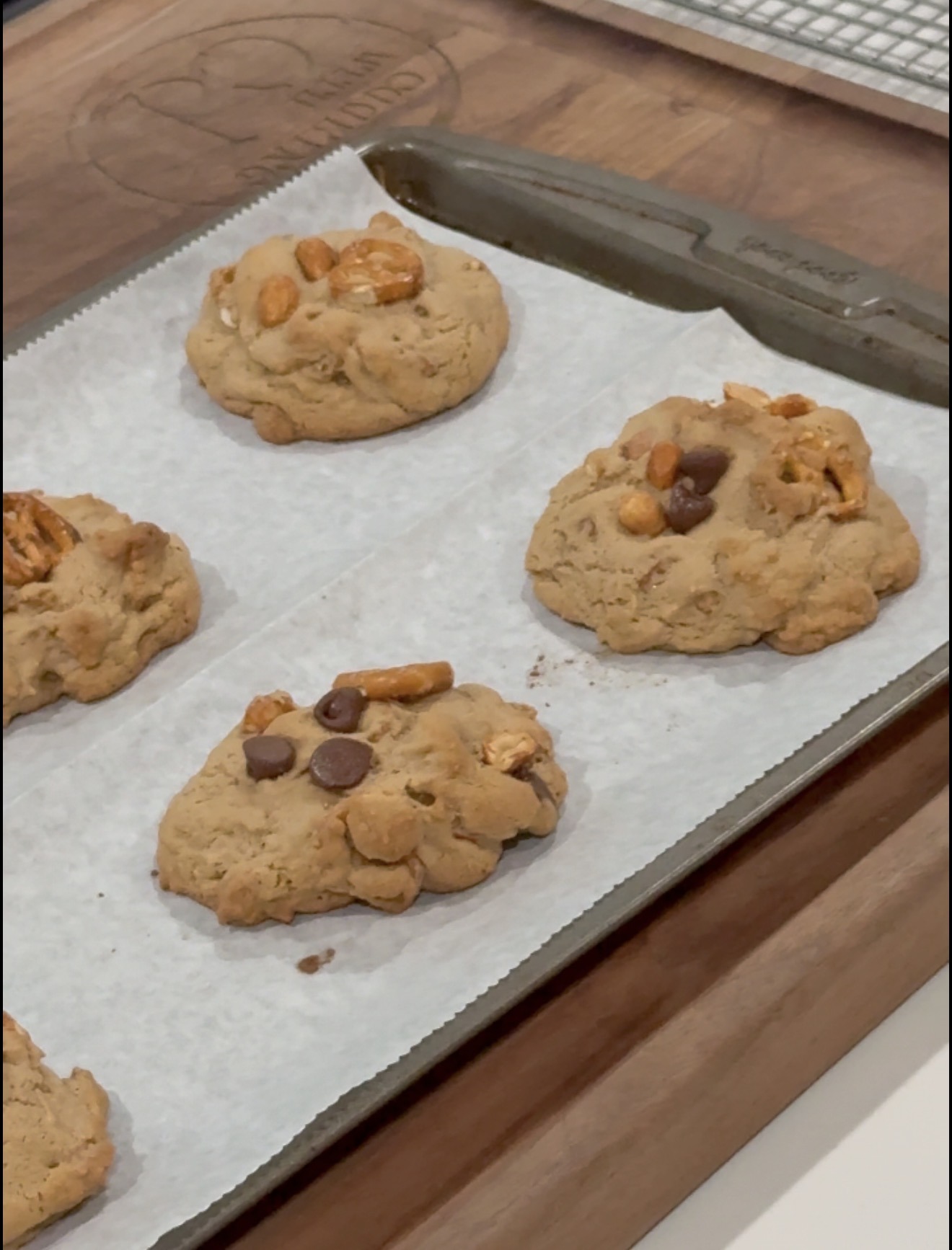 Five large cookies with chocolate chips, pretzel pieces, and nuts are on a baking sheet lined with parchment paper. The baking sheet rests on a wooden surface.