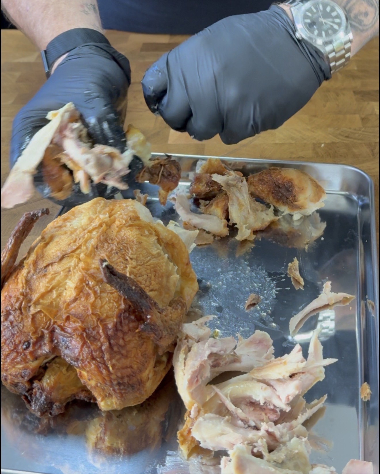 A person wearing black gloves shreds a roasted chicken on a metal tray, with pieces of chicken scattered around on a wooden surface.
