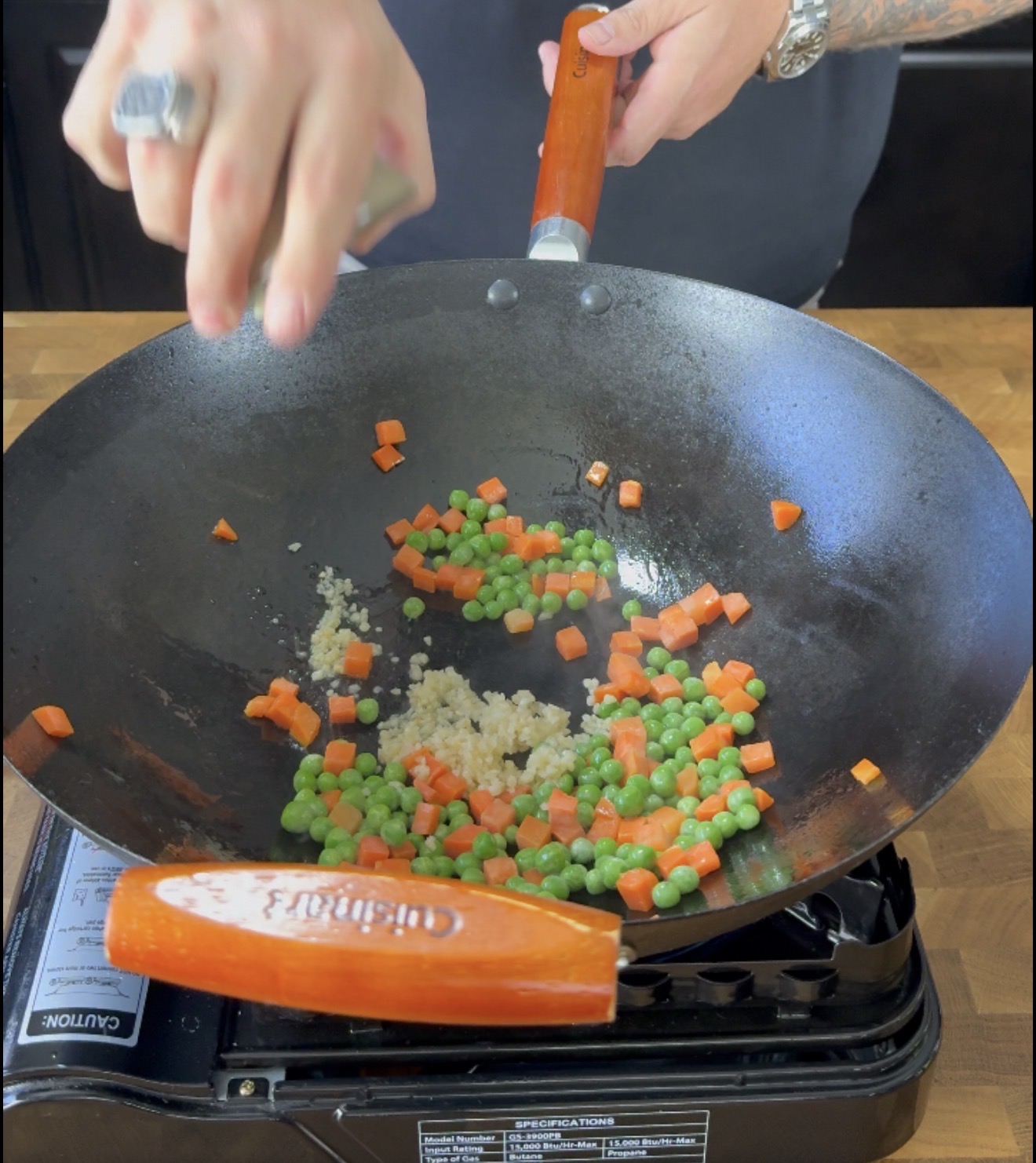 A hand stirs garlic, peas, and diced carrots in a black wok on a portable gas stove, with wooden handles visible.