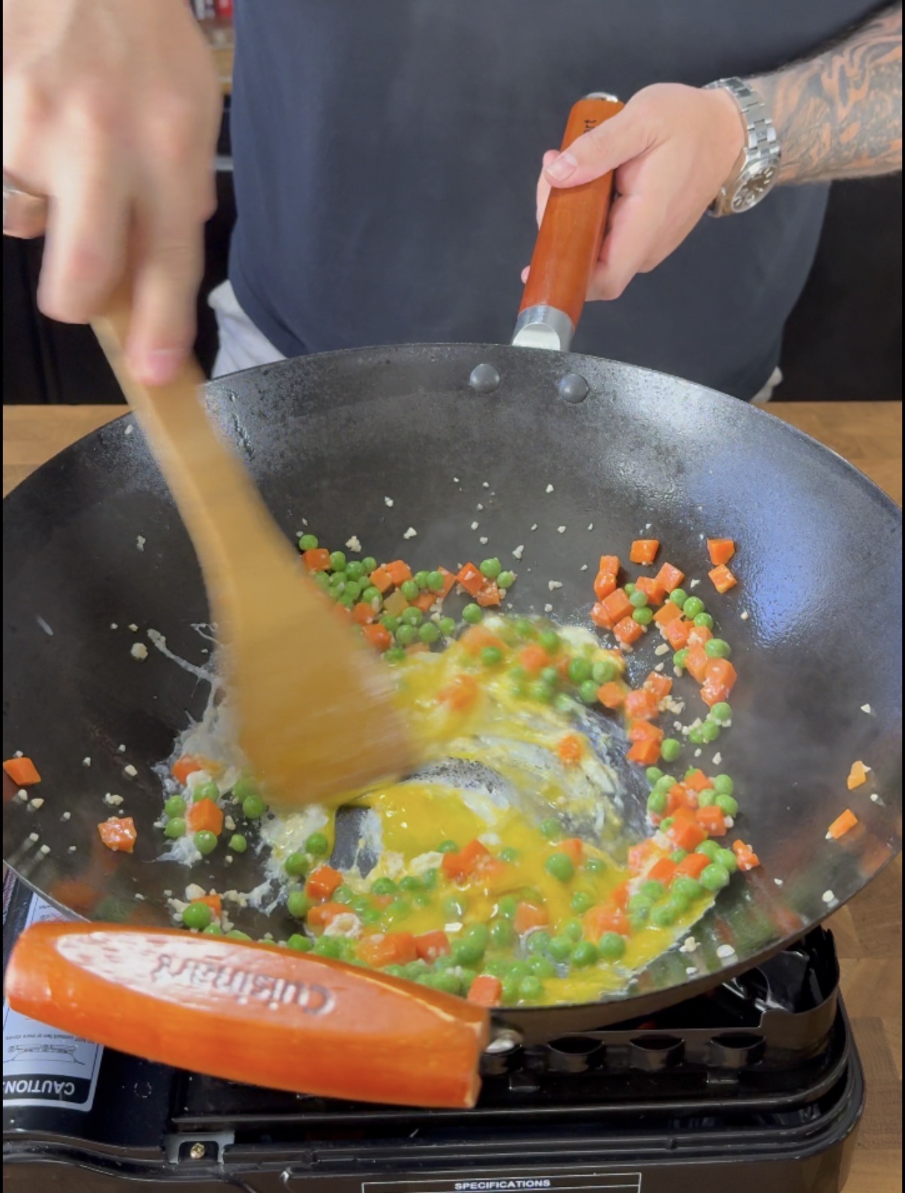 A person stirs eggs, peas, and diced carrots in a wok with a wooden spatula on a gas stove.