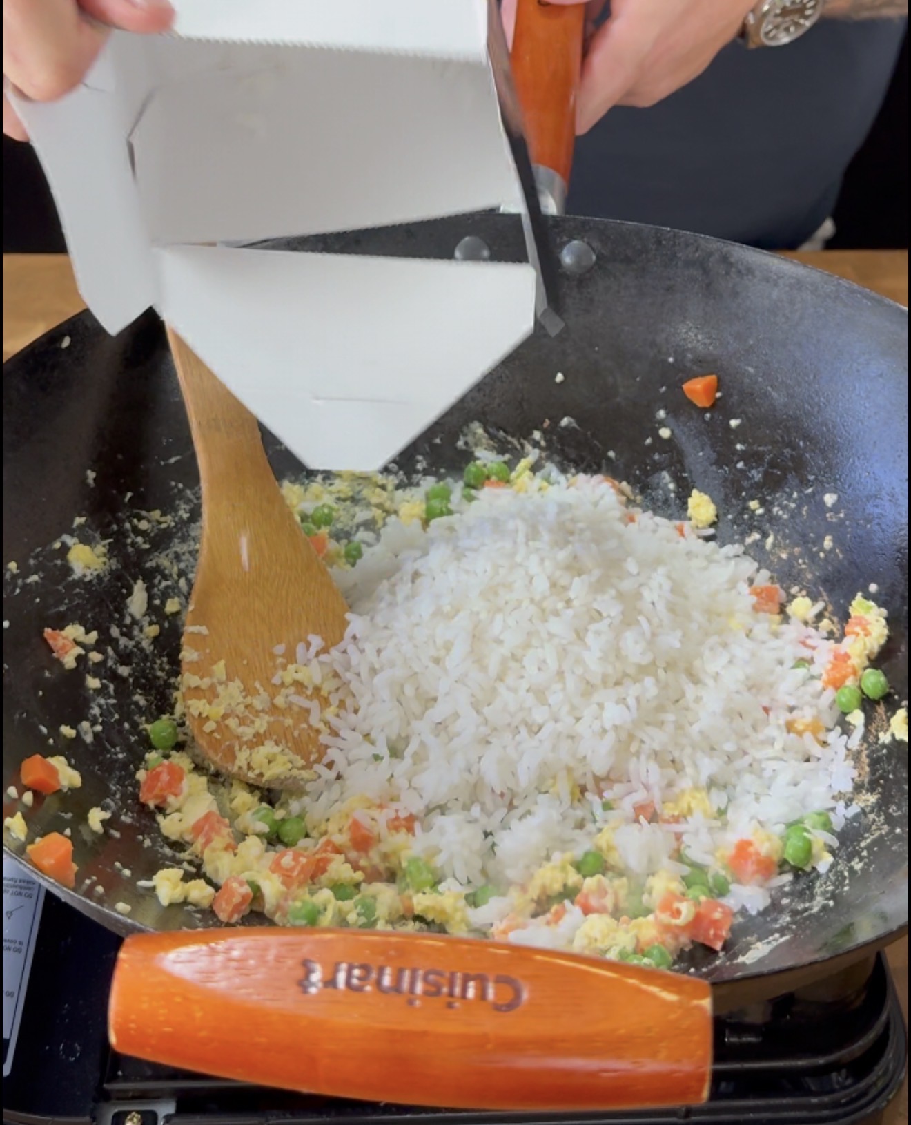 A person adds white rice from a carton into a wok containing mixed vegetables and scrambled eggs, stirring with a wooden spoon.