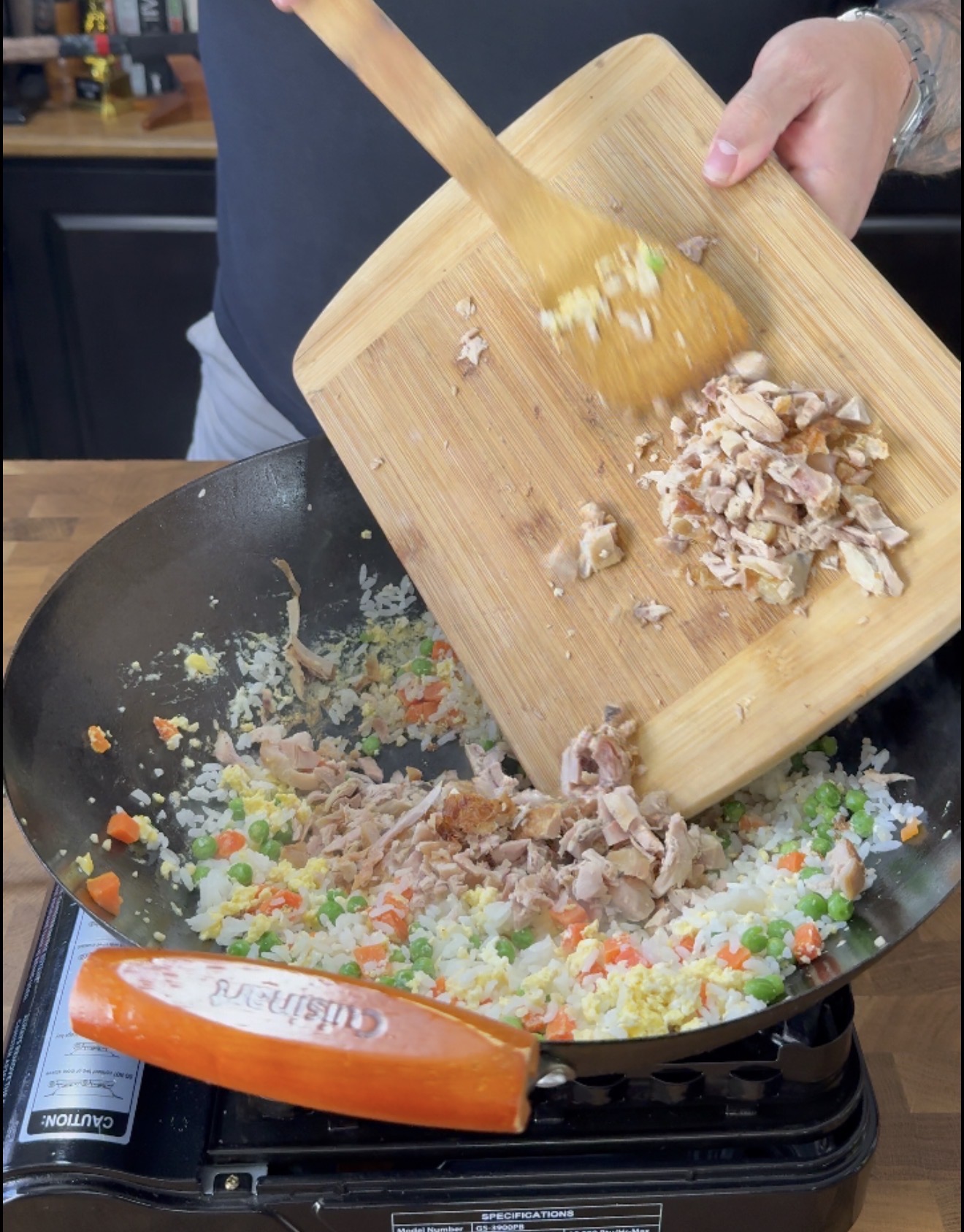 A person adds cooked chicken from a wooden cutting board into a wok with fried rice, peas, carrots, and egg on a stovetop.