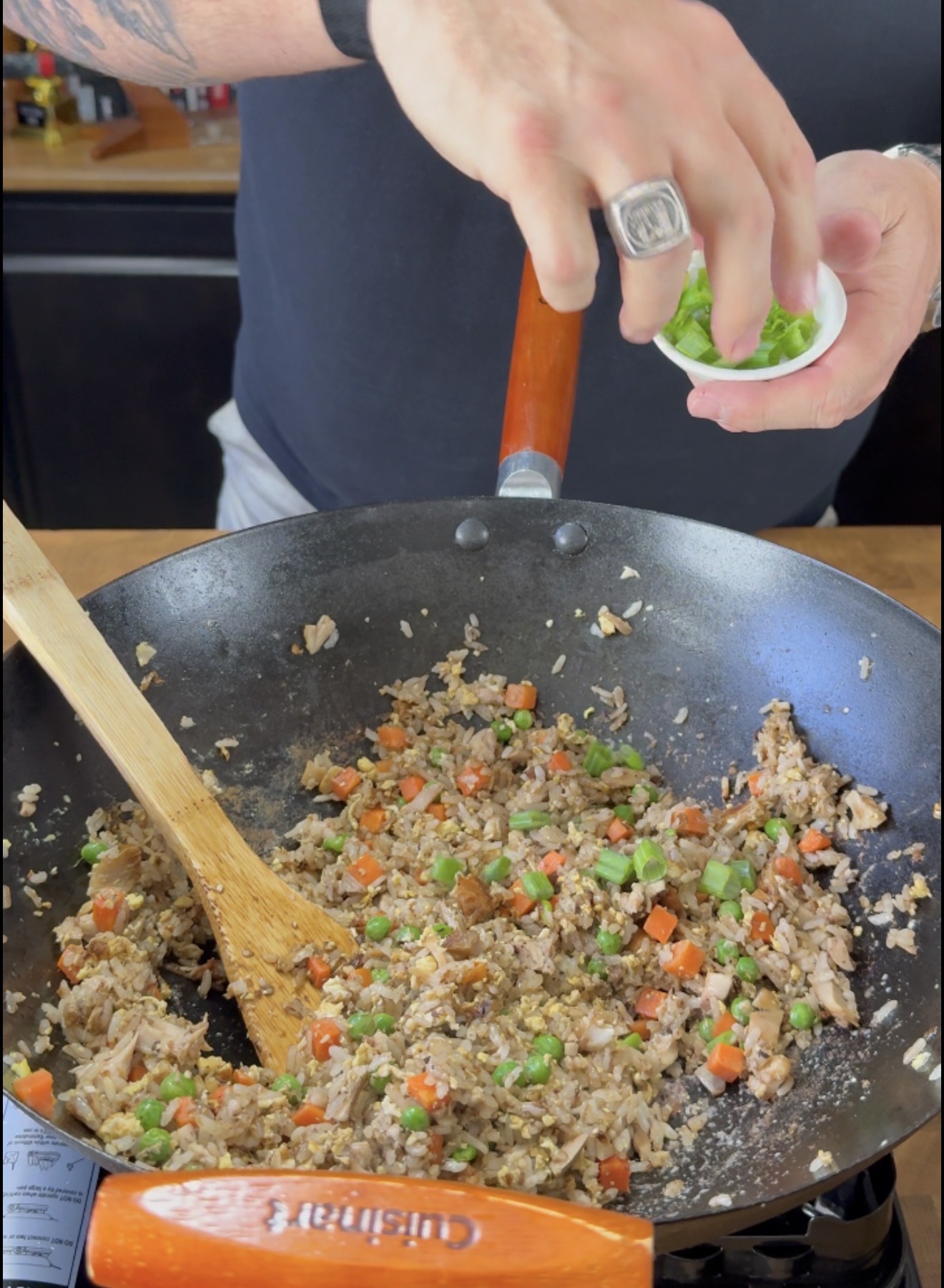 A person sprinkles chopped green onions into a wok filled with fried rice, diced carrots, and peas. A wooden spatula is in the pan, and the person is wearing a ring and a black shirt.