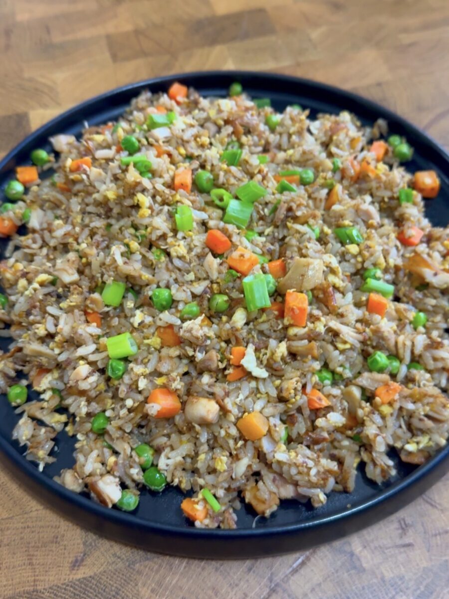 A black plate filled with fried rice mixed with green peas, diced carrots, chopped green onions, scrambled eggs, and small pieces of chicken sits on a wooden surface.