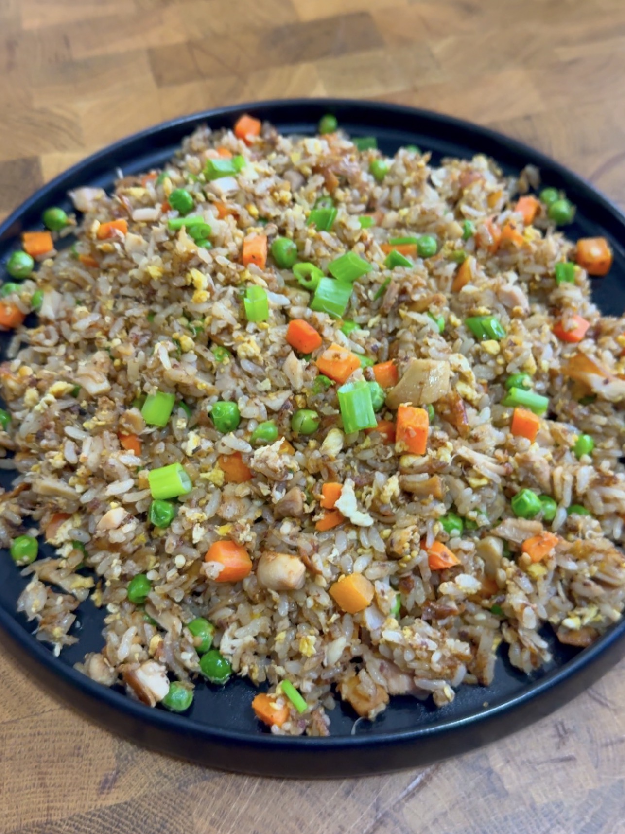 A black plate filled with fried rice mixed with green peas, diced carrots, chopped green onions, scrambled eggs, and small pieces of chicken sits on a wooden surface.