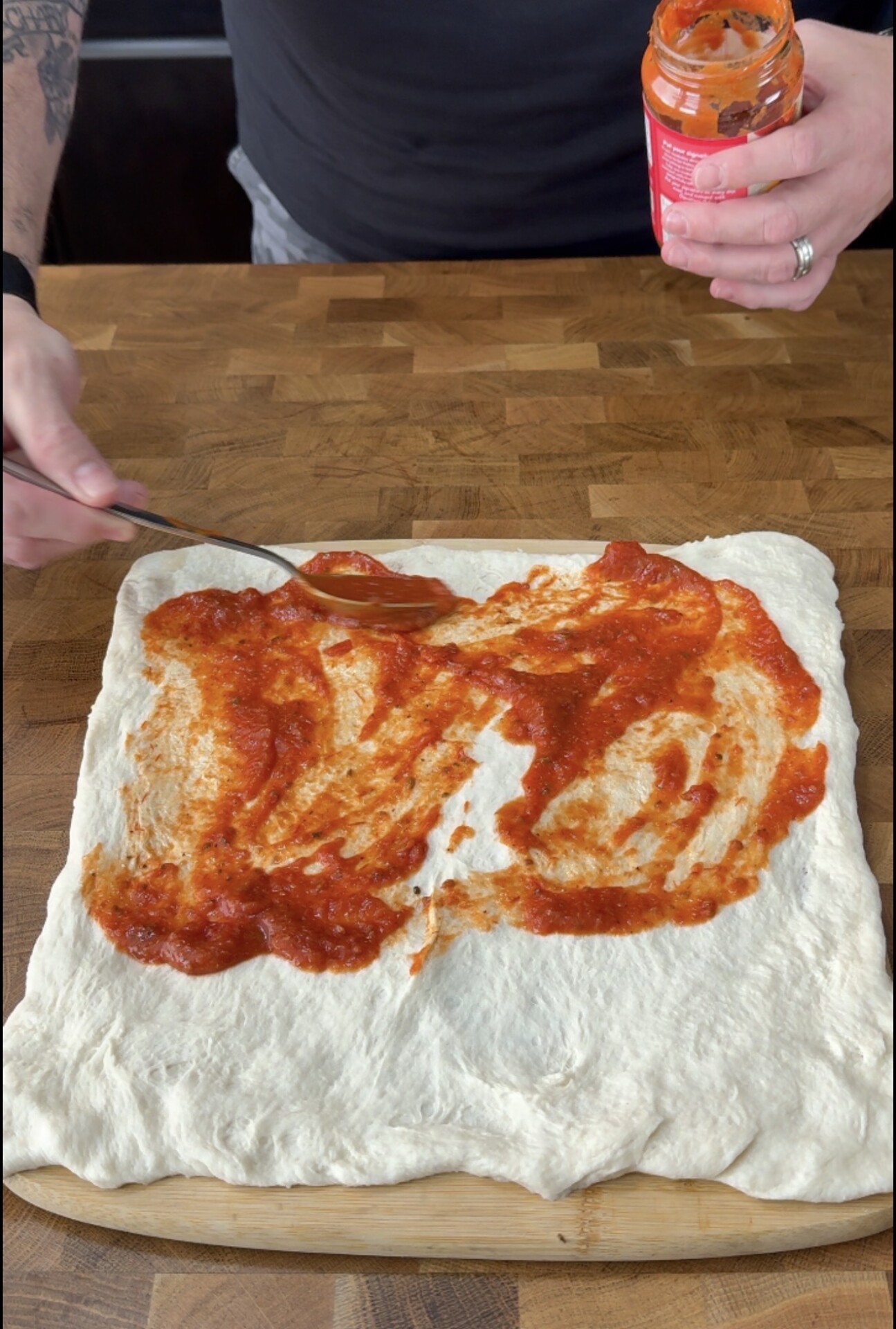 A person spreads tomato sauce on a rectangular pizza dough with a spoon on a wooden surface. A jar of sauce is held in the other hand.