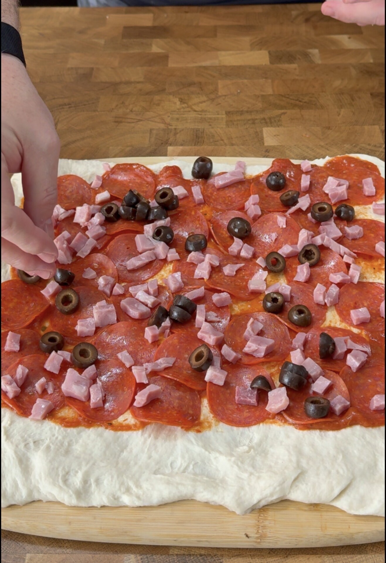 Hands adding toppings to a rectangular pizza dough covered with pepperoni, diced ham, black olives, and tomato sauce on a wooden surface.