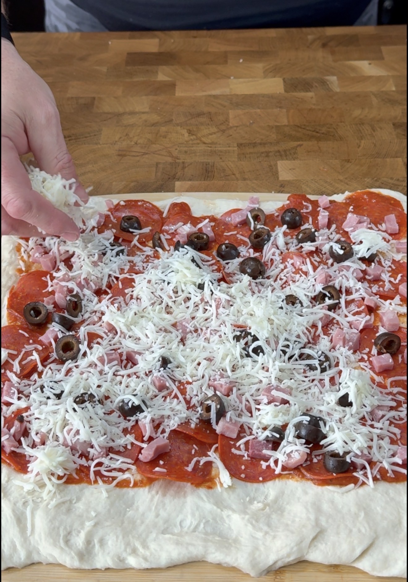 A hand sprinkles shredded cheese onto a rectangular pizza dough topped with pepperoni, olives, and ham on a wooden surface.