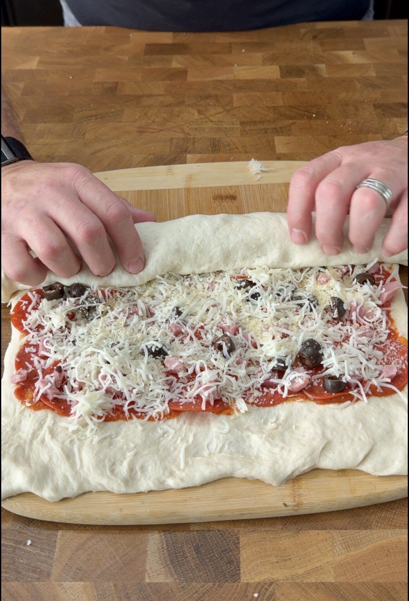 Hands roll up dough filled with tomato sauce, cheese, pepperoni, and black olives on a wooden surface, preparing a stuffed bread or pizza roll.