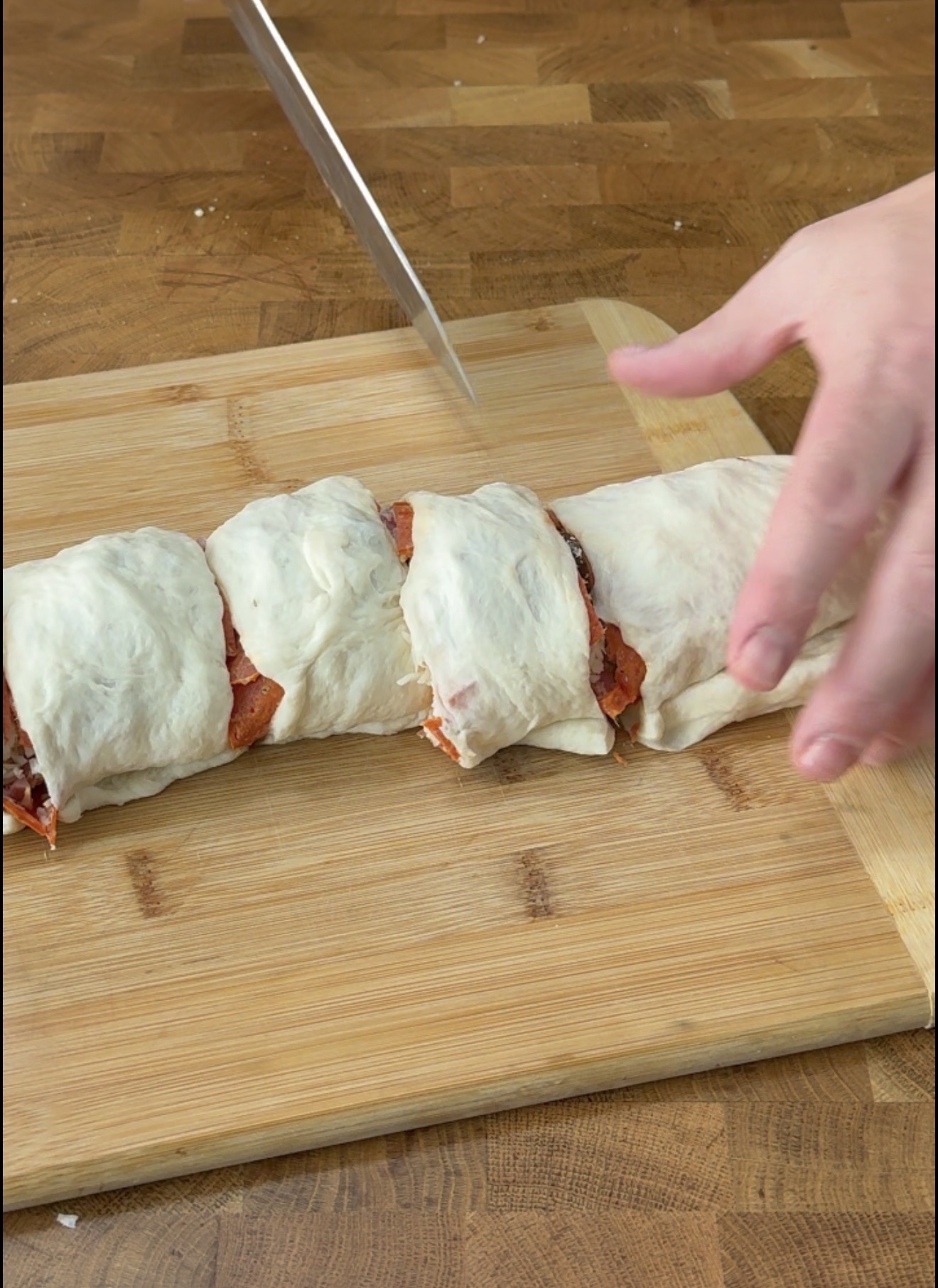 A hand slices a rolled dough with filling, possibly pizza, on a wooden cutting board with a knife.