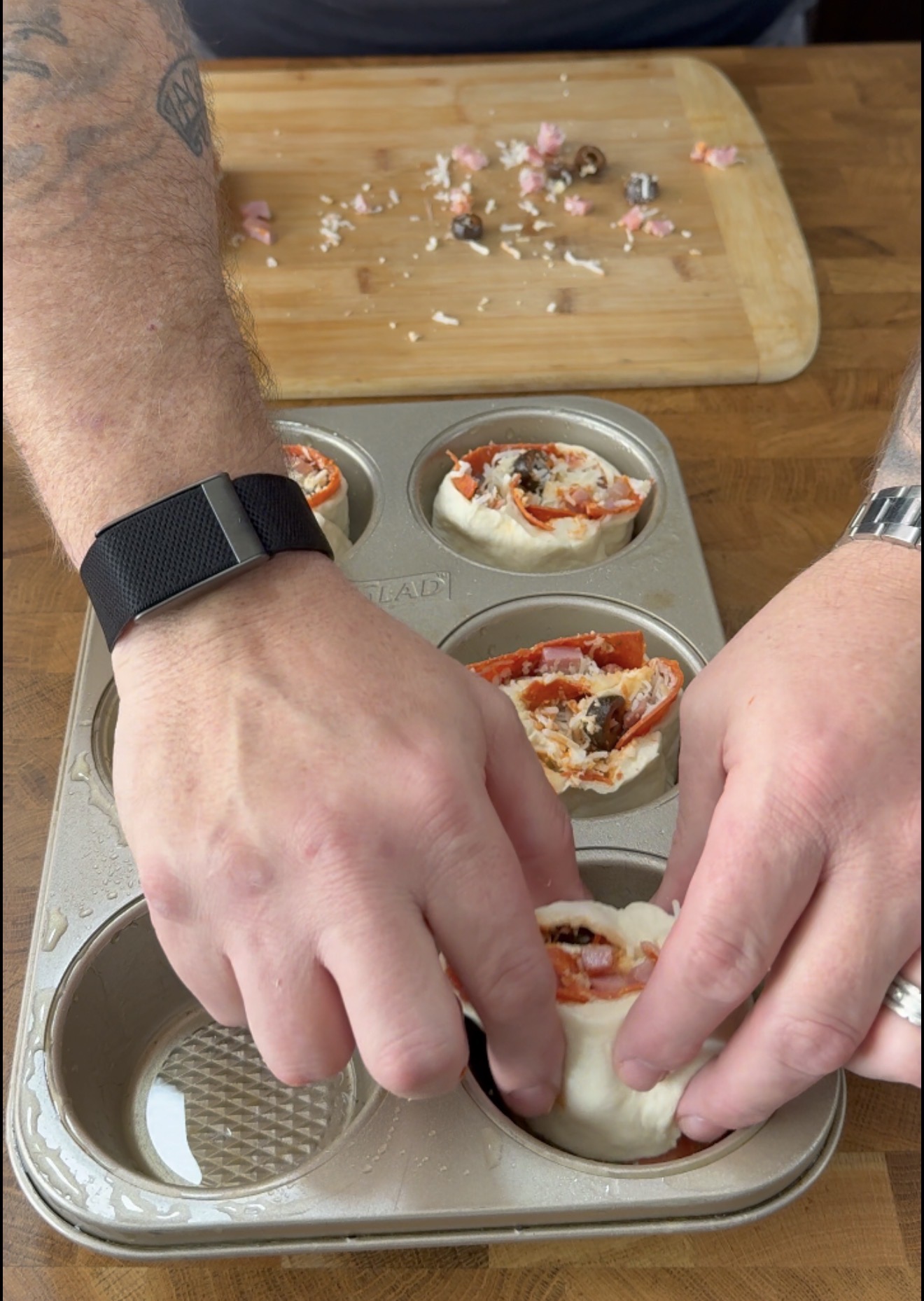 Close-up of hands placing rolled pizza dough with toppings into a muffin tin, with a cutting board and chopped ingredients in the background.