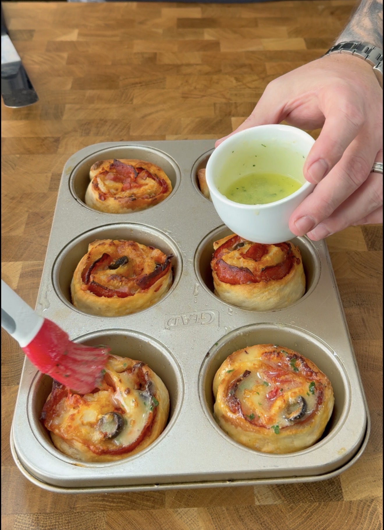 A hand brushes melted butter or sauce onto pizza rolls baked in a muffin tin. Another hand holds a small bowl of the sauce over the tin on a wooden countertop.