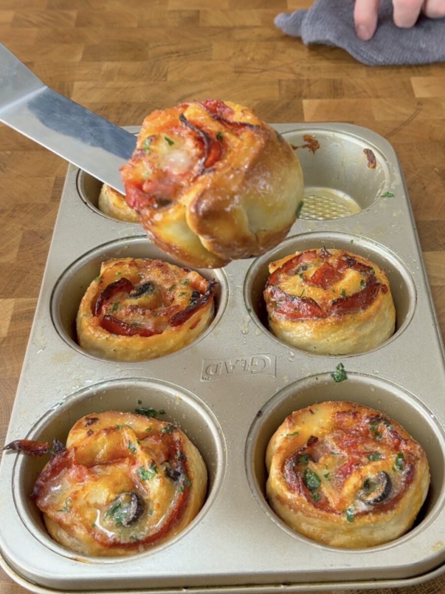 A spatula lifts a baked pizza roll from a muffin tin filled with other pizza rolls topped with cheese, pepperoni, and herbs.