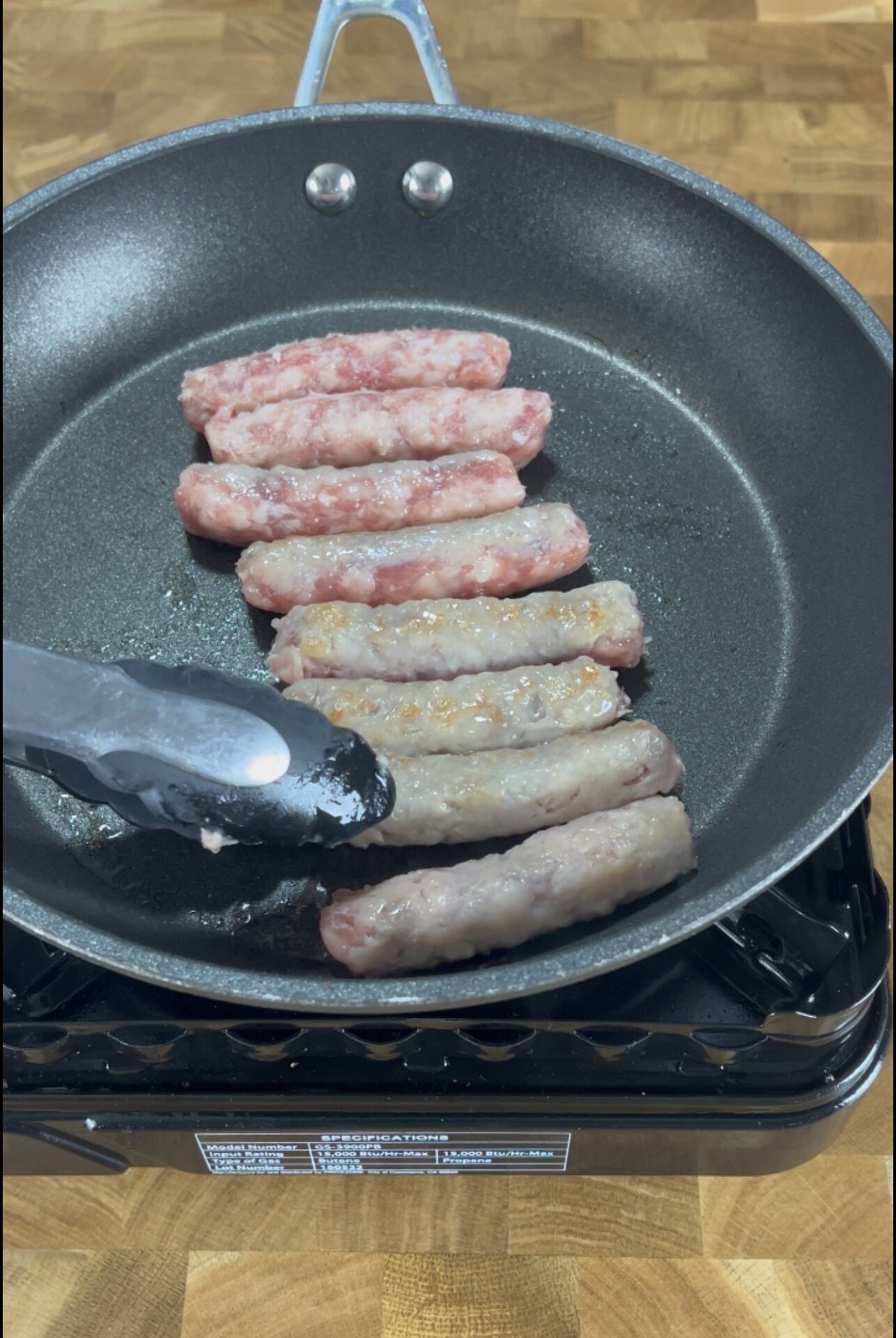 Seven sausage links are being cooked in a black frying pan on a stovetop. Some sausages appear more cooked than others. A pair of metal tongs is holding one sausage. The background is a wooden surface.
