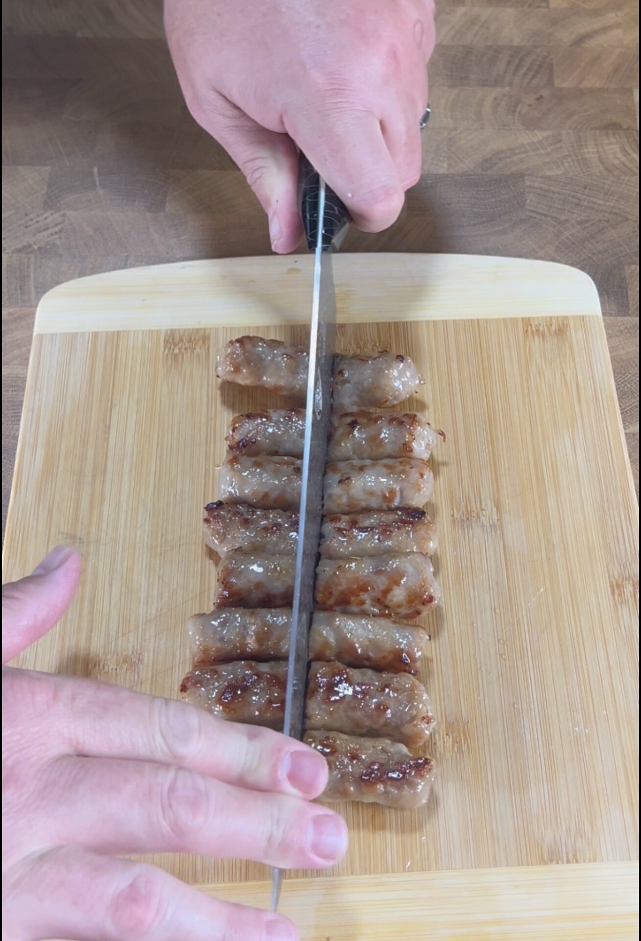 A hand slices cooked sausage links arranged in two neat rows on a wooden cutting board using a knife.