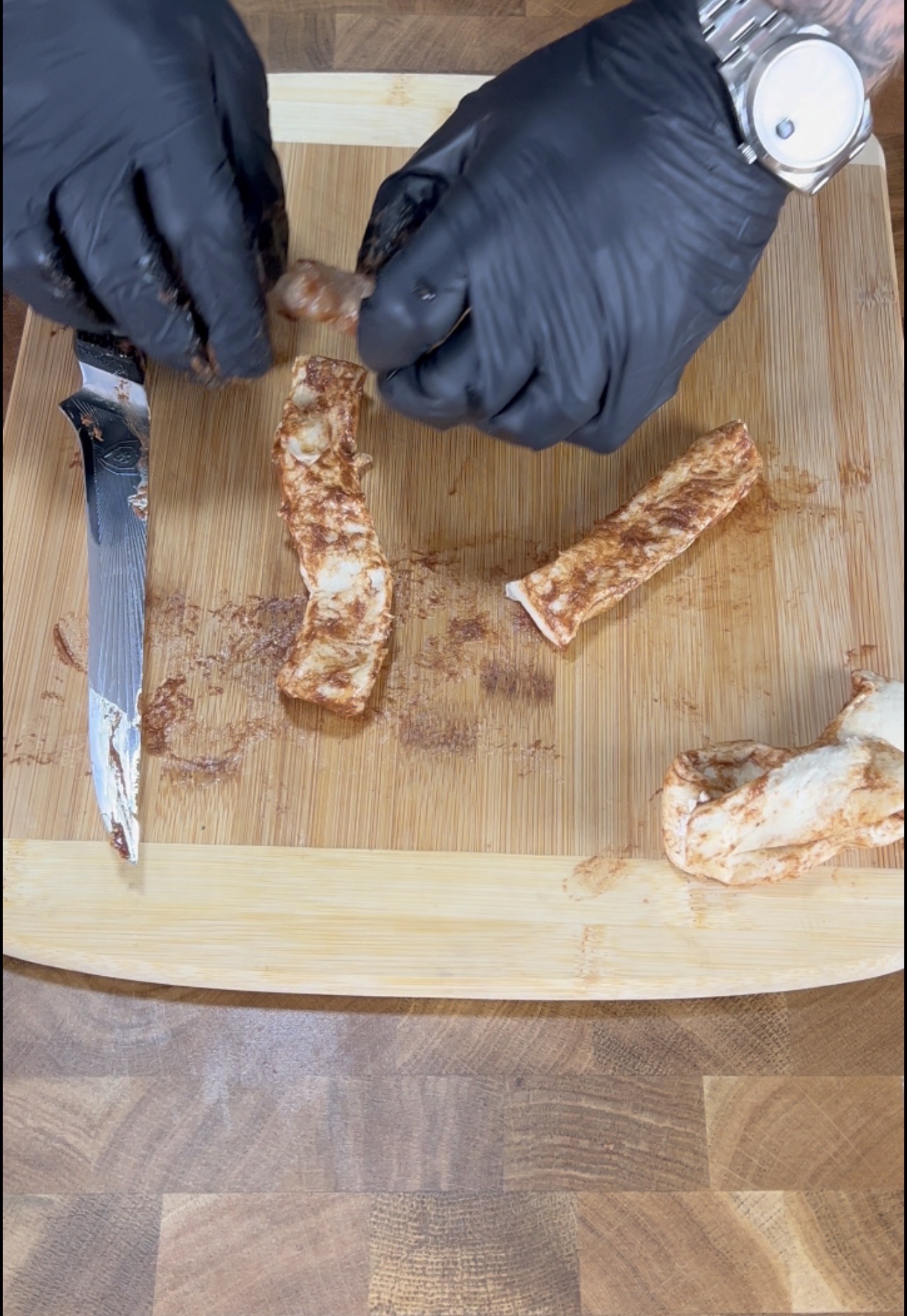 A person wearing black gloves slices a cinnamon roll on a wooden cutting board. A knife with cinnamon residue lies nearby, and three cut pieces of the cinnamon roll are visible on the board.