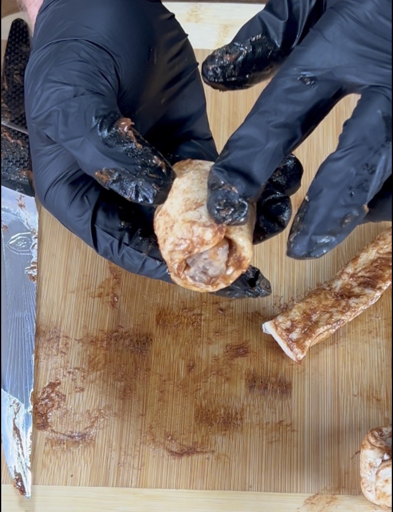 Gloved hands hold a seasoned raw meat piece above a wooden cutting board. A knife and other pieces of meat are visible, with seasoning scattered on the board.