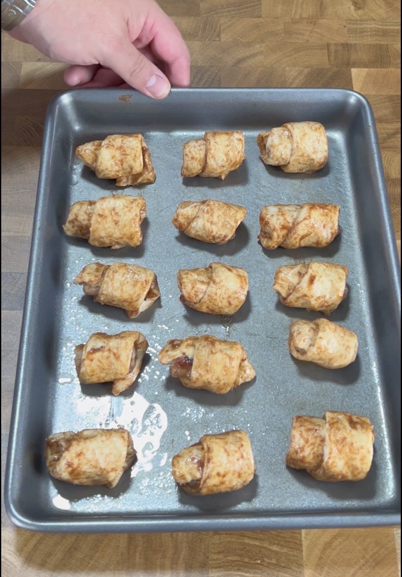 A hand holds a baking tray with fifteen small, golden-brown pastry rolls spaced evenly on it, resting on a wooden surface.