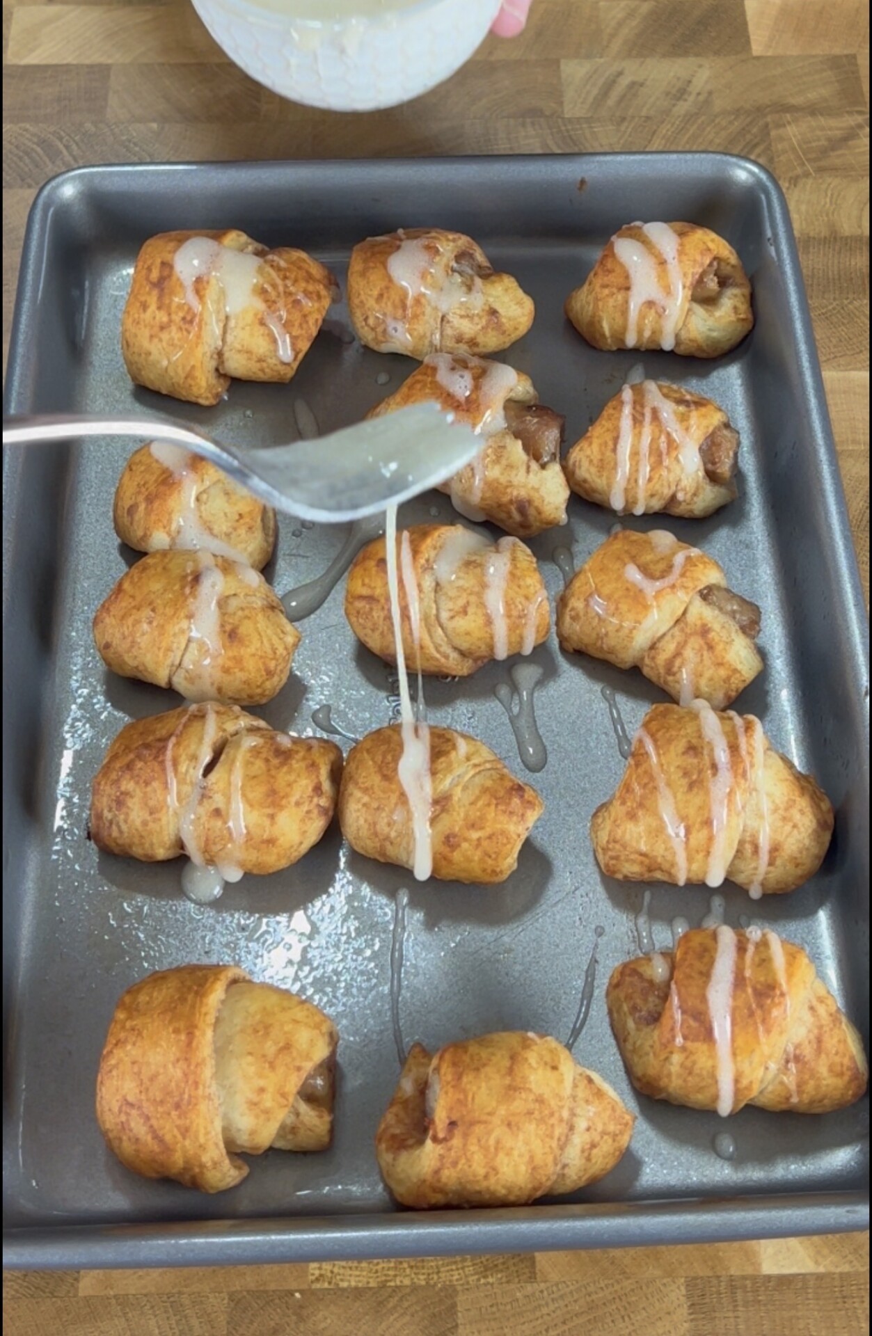 A spoon drizzles white icing over golden-brown crescent roll pastries filled with apple pieces, arranged on a baking sheet atop a wooden surface.