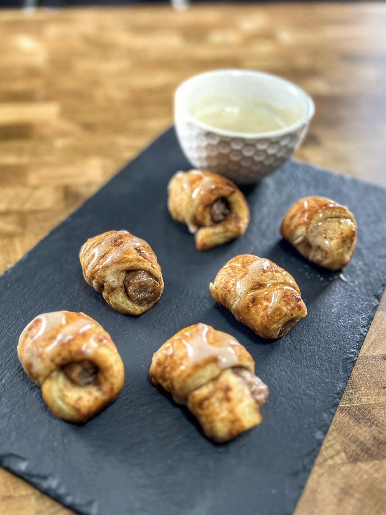Six golden-brown pastry rolls are arranged on a black slate, with a small white bowl of icing glaze in the background on a wooden table.