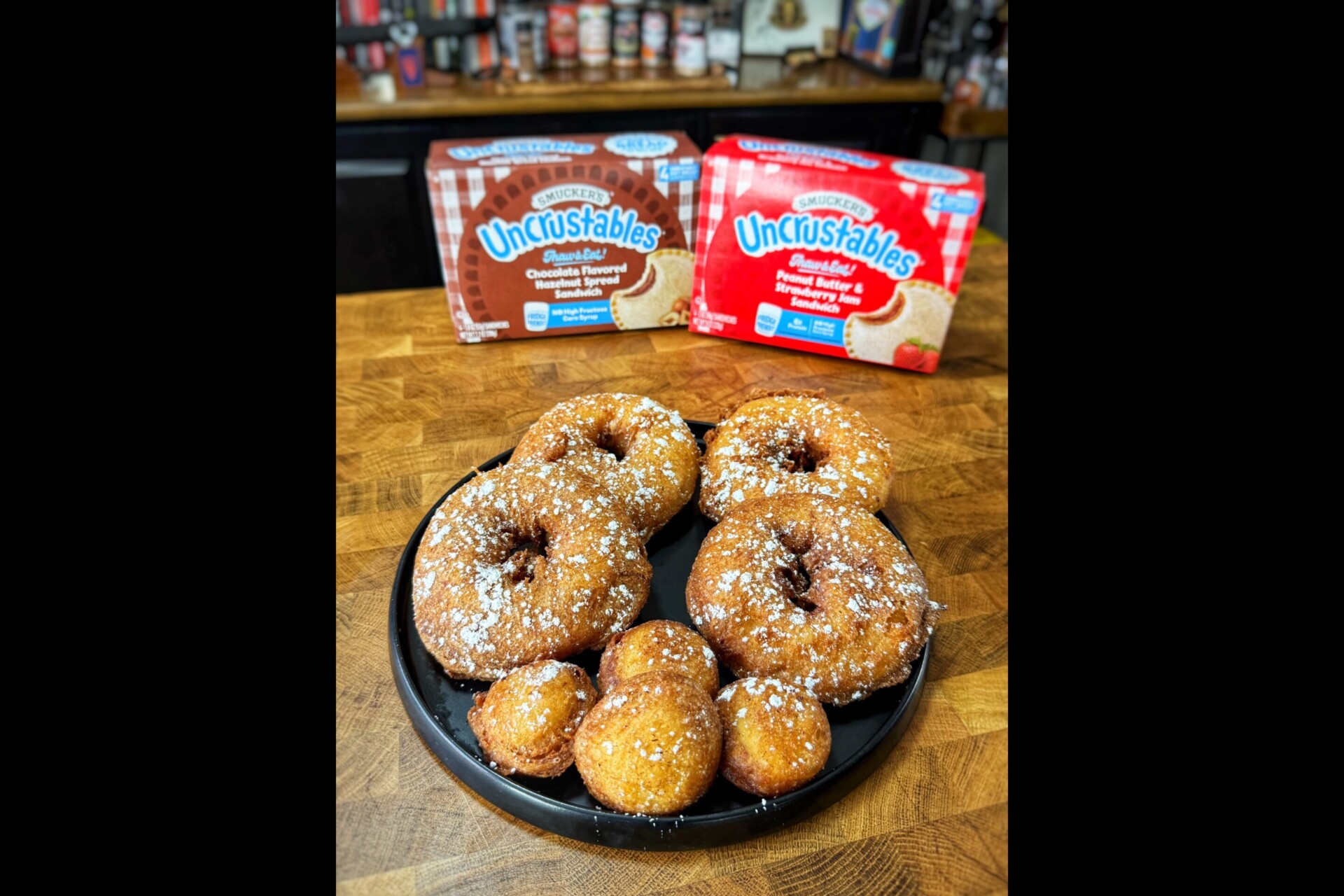 A plate of deep-fried Uncrustables sandwiches and balls is dusted with powdered sugar. Two Uncrustables packages, one strawberry and one peanut butter and grape jelly, are in the background on a wooden counter.