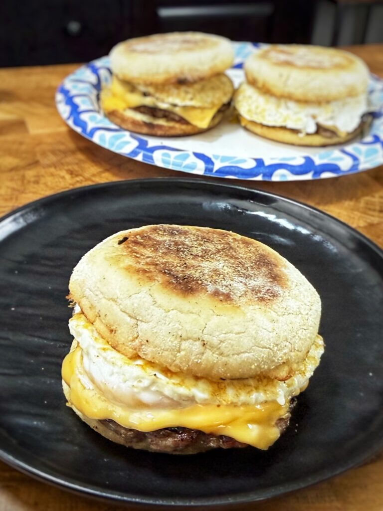 A breakfast sandwich with egg, cheese, and sausage on an English muffin sits on a black plate, with two more sandwiches on a blue and white plate in the background.