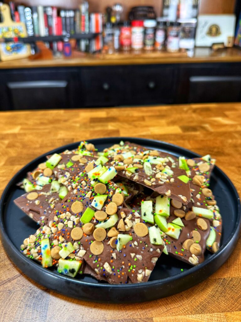 A black plate holds pieces of chocolate bark topped with green apple slices, sprinkles, chopped nuts, and light brown candy, on a wooden counter with shelves and spices blurred in the background.