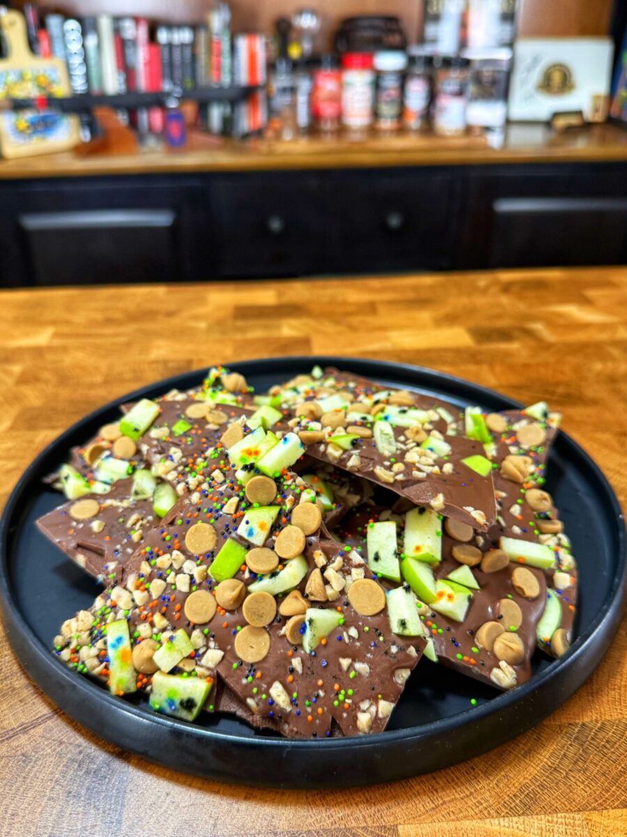 A black plate holds pieces of chocolate bark topped with green apple slices, sprinkles, chopped nuts, and light brown candy, on a wooden counter with shelves and spices blurred in the background.
