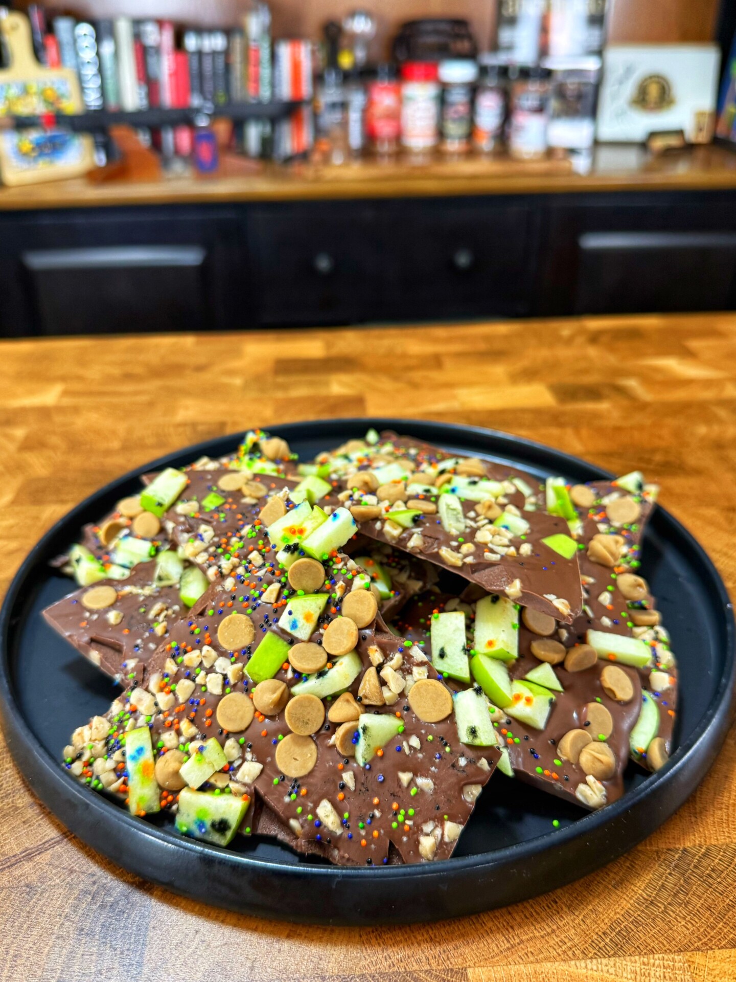 A black plate holds pieces of chocolate bark topped with green apple slices, sprinkles, chopped nuts, and light brown candy, on a wooden counter with shelves and spices blurred in the background.
