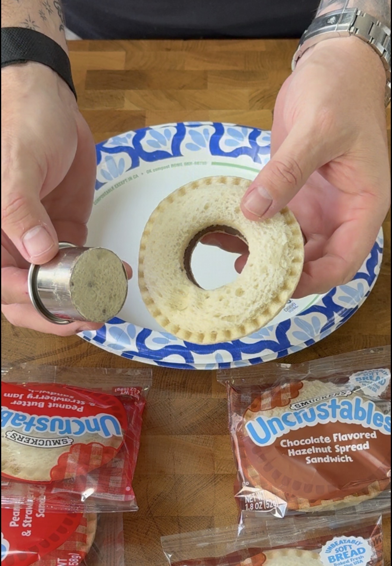 A person holds a metal cutter and a round sandwich with the center removed, over a paper plate. Uncrustables chocolate hazelnut sandwiches are on the table.