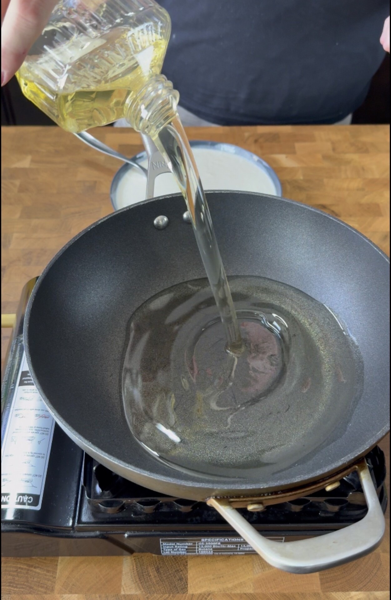 A person pours cooking oil from a bottle into a black frying pan on a portable stovetop, with a wooden counter in the background.