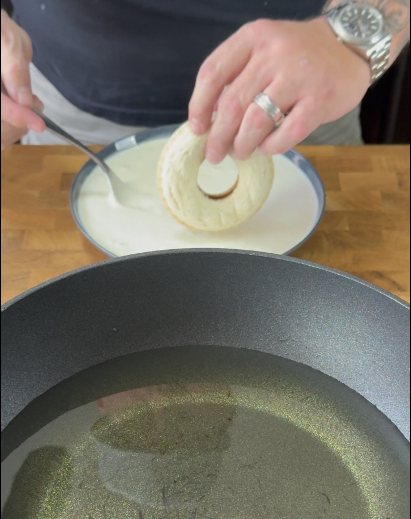 A person dips a round slice of bread with a hole in the center into a plate of white batter, preparing it for frying in hot oil in a black pan on a wooden countertop.