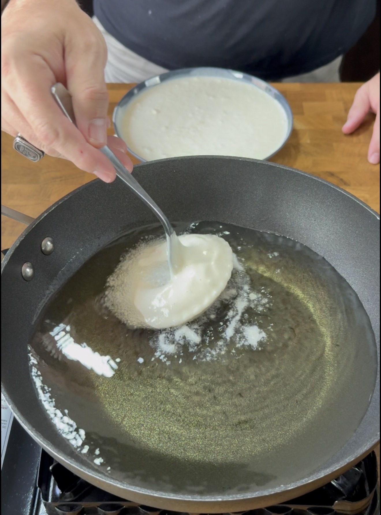 A person uses a spoon to place batter into a pan of hot oil for frying. A plate with more batter is visible in the background on a wooden surface.
