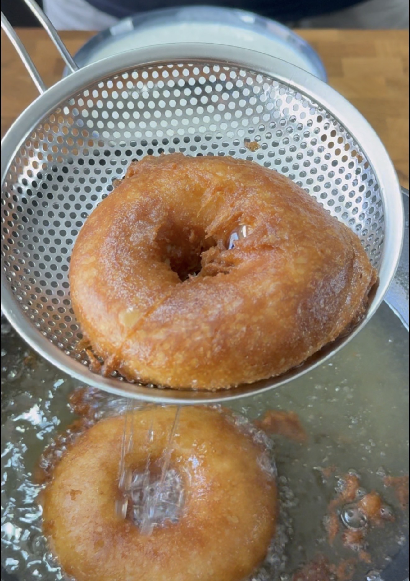 A golden-brown donut is being lifted from hot oil with a metal slotted spoon, while another donut fries below in the bubbling oil.